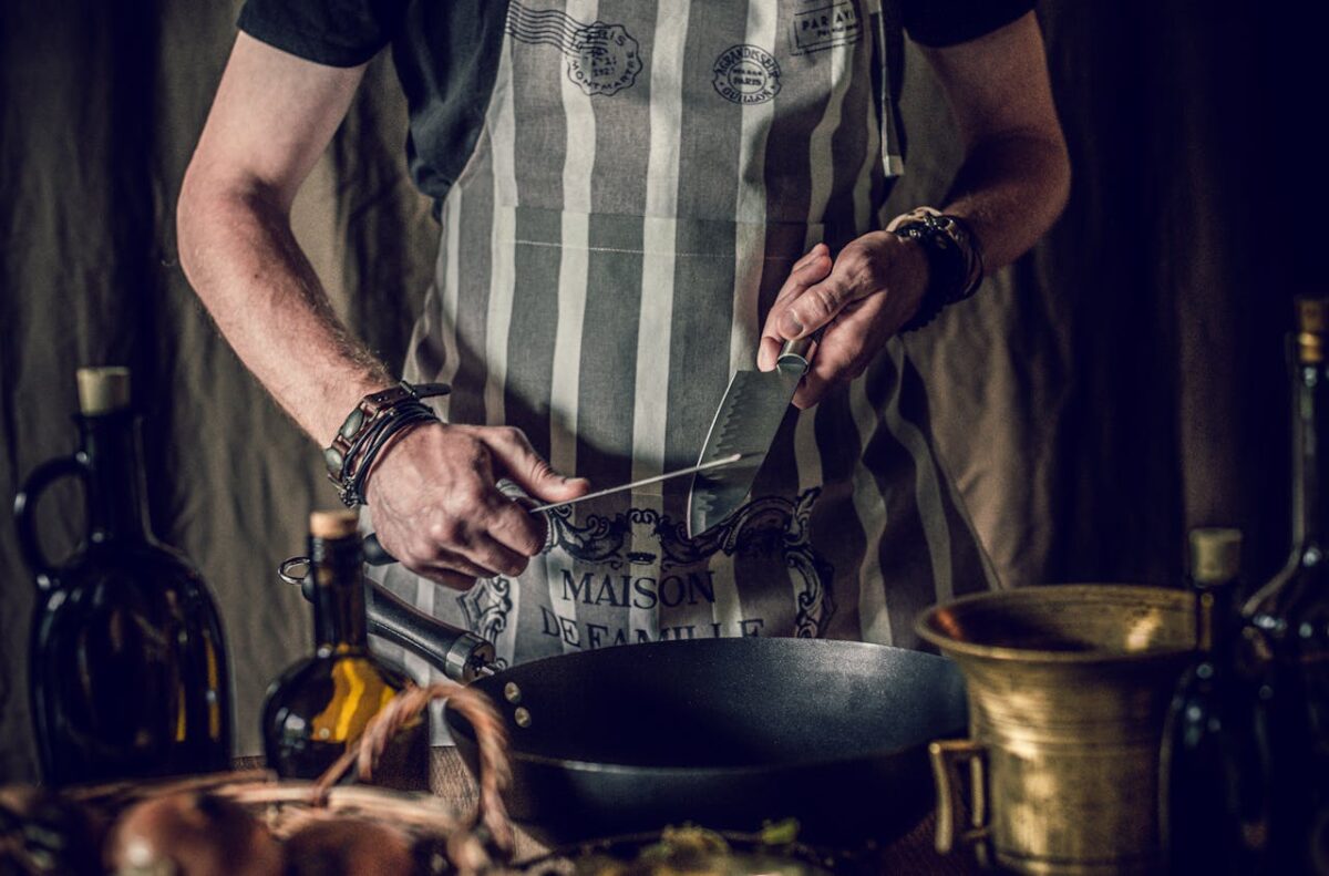 Crop anonymous male cook in apron holding sharp knives in hands while standing at table with frying pan and oil bottles and preparing lunch